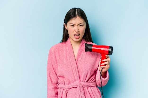 Jovem mulher asiática vestindo um roupão de banho e segurando secador de cabelo isolado no fundo rosa gritando muito irritado e agressivo