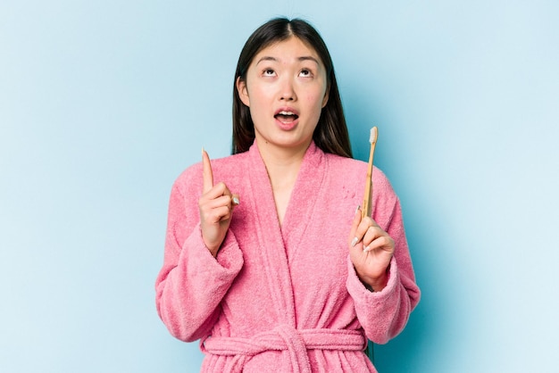 Jovem mulher asiática vestindo um roupão de banho escovando os dentes isolados no fundo rosa apontando para cima com a boca aberta