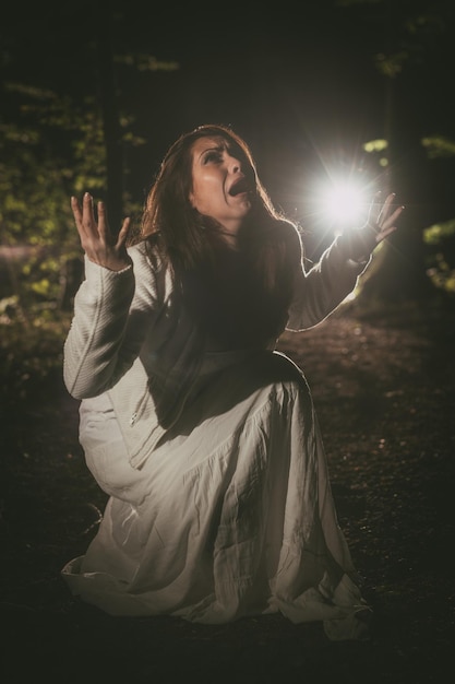 Jovem mulher assustada gritando na floresta à noite em vestido branco.