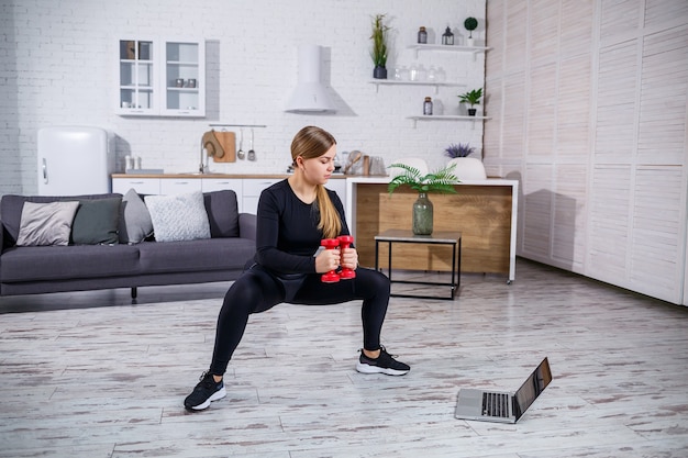 Jovem mulher atlética em roupas de fitness, blusa preta e leggings em um apartamento, usando exercícios online de um site de fitness em um laptop e praticando esportes em casa. fitness em casa durante a quarentena