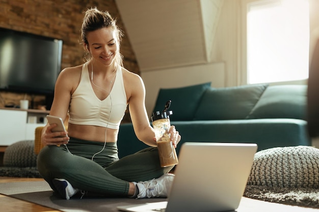 Jovem mulher atlética feliz relaxando no chão e usando laptop enquanto bebe smoothie em casa