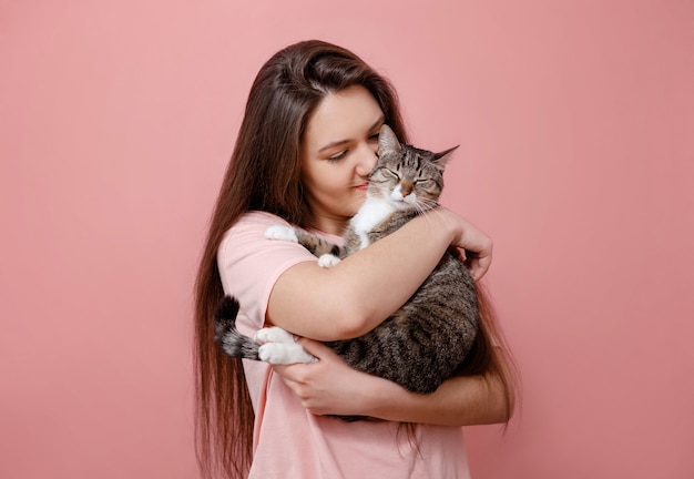 Jovem mulher atraente abraçando o gato nas mãos, fundo rosa, menina beijando o gato