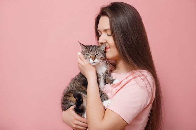 Foto jovem mulher atraente abraçando o gato nas mãos, fundo rosa