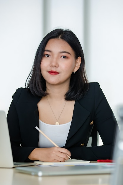 Jovem mulher atraente asiática em um terno preto sentado no escritório moderno, trabalhando e sorrindo com o laptop em cima da mesa. Conceito de estilo de vida de escritório moderno.
