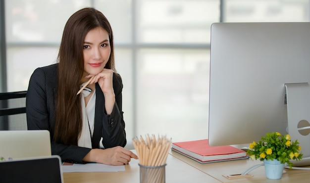 Jovem mulher atraente asiática no negócio negro, sentado atrás da mesa com o computador e o laptop no escritório moderno com fundo borrado do windows. Conceito de estilo de vida de escritório moderno.