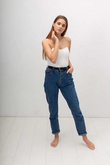 Jovem mulher atraente caucasiana com cabelos castanhos compridos em calça jeans espartilho, azul sobre fundo branco do estúdio. senhora bonita magra posando com os pés descalços na sala iluminada. retrato de linda mulher