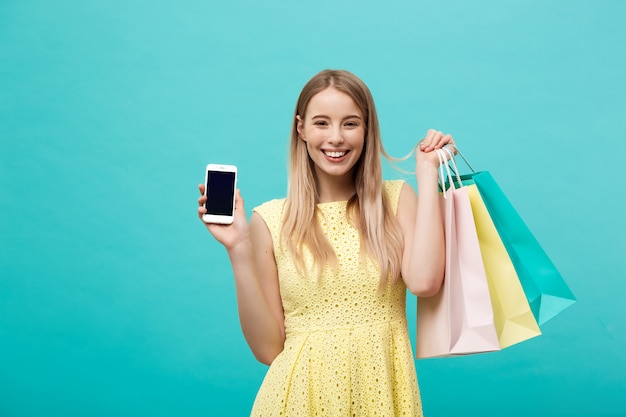 Jovem mulher atraente com sacos de compras mostra a tela do telefone diretamente para a câmera.