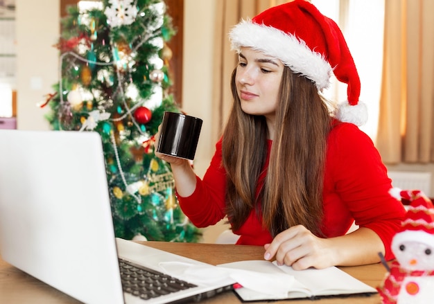 Jovem mulher atraente com suéter vermelho e chapéu de papai noel trabalhando em casa durante as férias de natal.