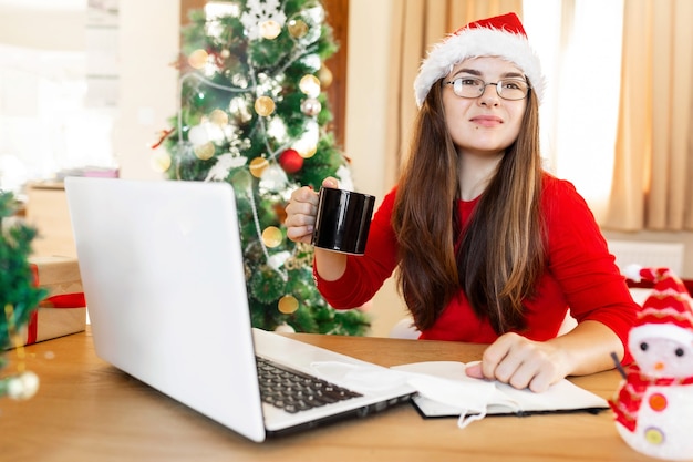 Foto jovem mulher atraente com suéter vermelho e chapéu de papai noel trabalhando em casa durante as férias de natal.