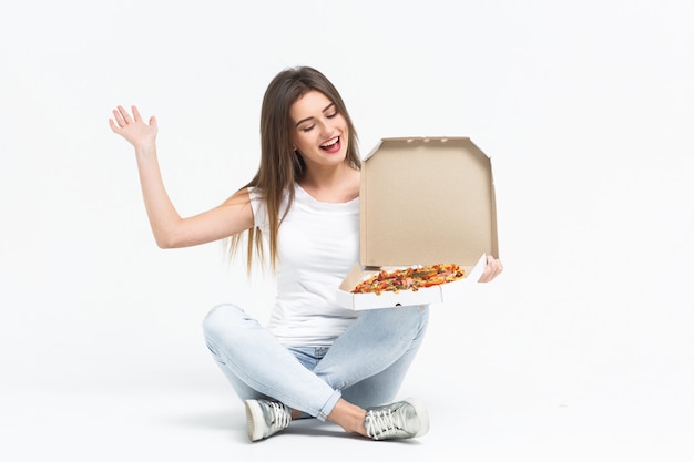 Jovem mulher atraente, comer um pedaço de pizza deliciosa. Ela é uma camiseta, jeans e tênis sentados no chão em casa. Entrega de alimentos.