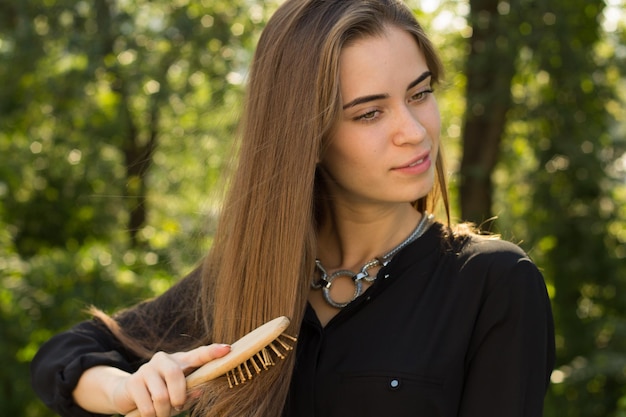 Jovem mulher atraente de blusa preta e colar prateado penteando seu cabelo comprido castanho no fundo de árvores verdes