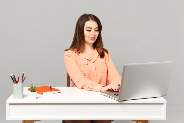 Jovem mulher atraente em roupas casuais pastel senta-se, trabalha na mesa branca com laptop pc isolado em fundo cinza, retrato de estúdio. Conceito de estilo de vida de carreira de negócios de realização. Mock up espaço de cópia.