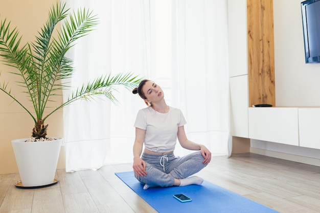 Jovem mulher atraente está praticando ioga online usando um aplicativo de celular ou smartphone. Manhã de ioga feminina com roupas casuais em casa, na sala de estar, posição de lótus de treino meditando em uma esteira