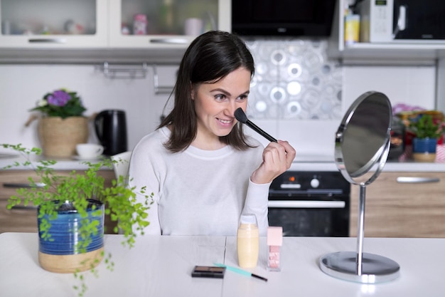 Jovem mulher atraente fazendo maquiagem, mulher sentada em casa à mesa da cozinha com escova de espelho de produtos cosméticos. Beleza, cuidados com a pele, moda, conceito de maquiagem