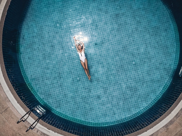 Jovem mulher atraente flutua nas costas na piscina azul redonda, senhora mantém os braços para cima, manchas na água clara. Conceito de recreação de luxo, vista aérea