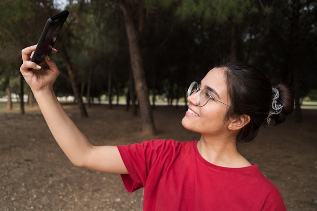 Jovem mulher atraente na casa dos vinte com óculos e uma camisa vermelha, sentado em um banco, segurando um telefone celular e tirar uma selfie enquanto sorrindo em um belo parque na espanha. ela está olhando o telefone