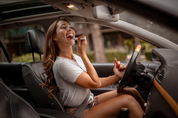 Foto jovem mulher atraente olhando no espelho retrovisor pintando os lábios fazendo aplicação de maquiagem no carro.