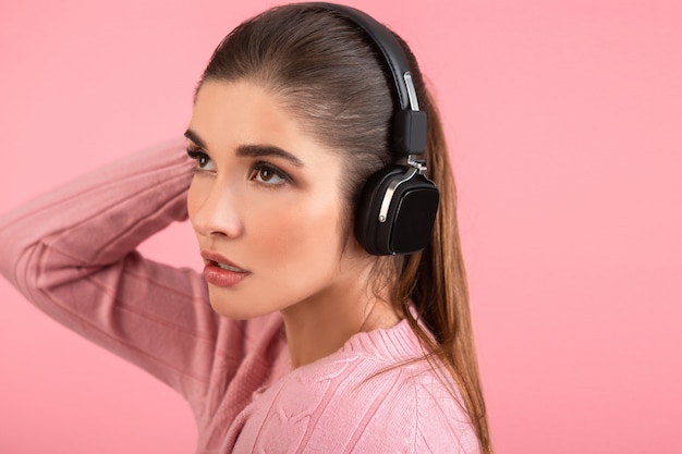 Jovem mulher atraente ouvindo música em fones de ouvido sem fio, vestindo um suéter rosa, sorrindo, feliz humor positivo, posando em fundo rosa