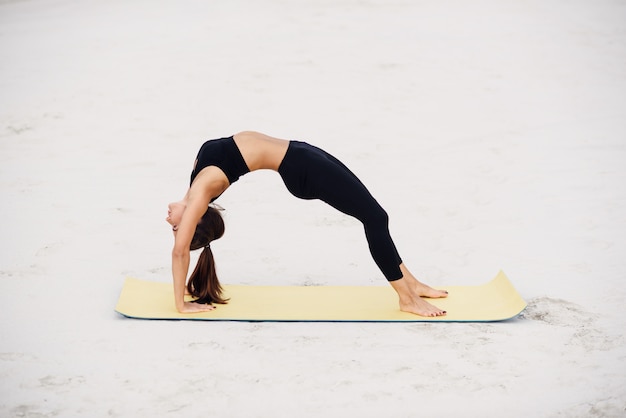 Jovem mulher atraente praticando ioga, estendendo-se no exercício de ponte. Malhando na praia. Ioga de esporte fitness e conceito de estilo de vida saudável.