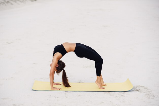 Jovem mulher atraente praticando ioga, estendendo-se no exercício de ponte. Malhando na praia. Ioga de esporte fitness e conceito de estilo de vida saudável.