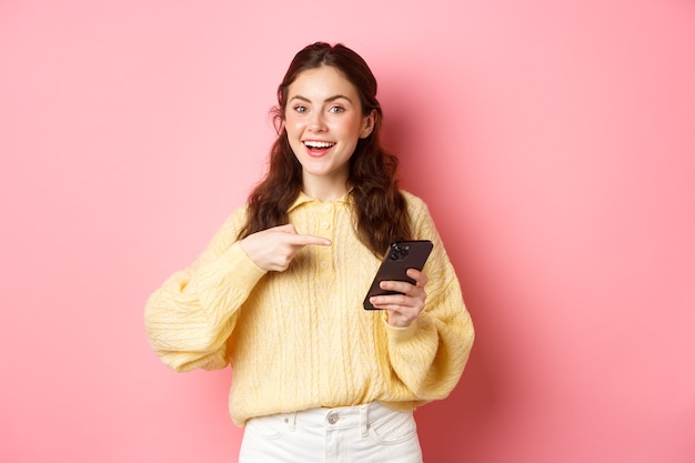 Jovem mulher atraente segurando o smartphone, apontando para a tela, promovendo, falando sobre sua página de mídia social, em pé sobre a parede rosa.