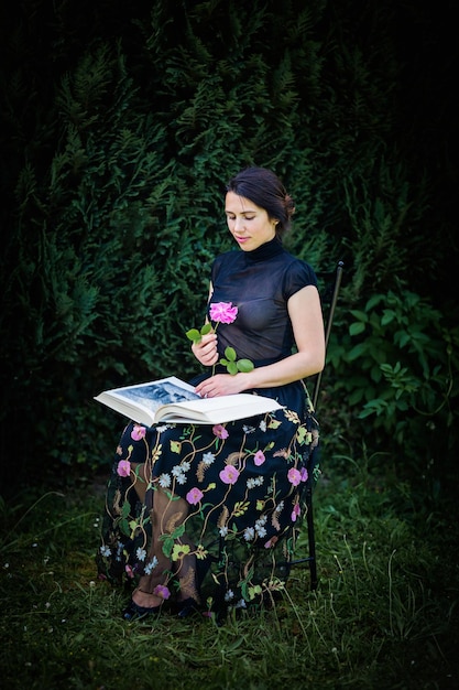 Jovem mulher atraente sentada em um jardim com um livro grande e uma flor rosa nas mãos