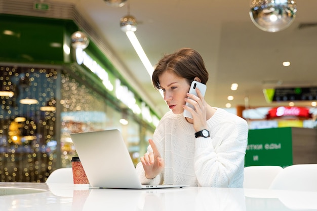 Jovem mulher atraente sentada em um shopping center em uma mesa e trabalhando em um laptop de computador