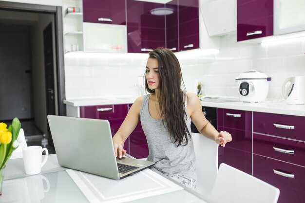 Jovem mulher atraente, trabalhando no laptop na cozinha. Fêmea, digitando no pc dentro de casa pela manhã.