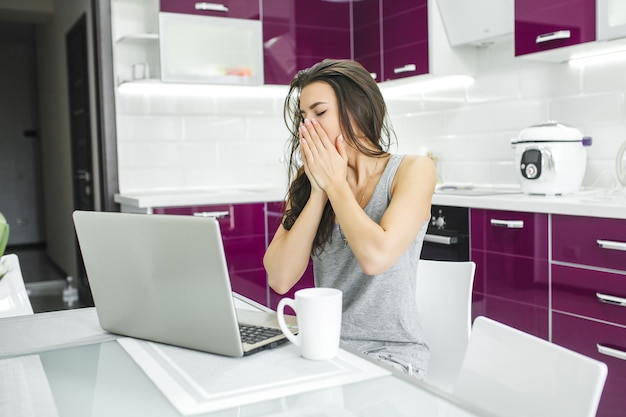 Jovem mulher atraente, trabalhando no laptop na cozinha. Fêmea, digitando no pc dentro de casa pela manhã.