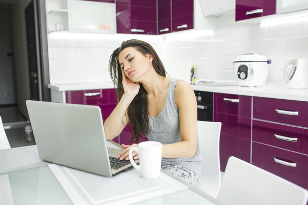 Jovem mulher atraente, trabalhando no laptop na cozinha. Fêmea, digitando no pc dentro de casa pela manhã.