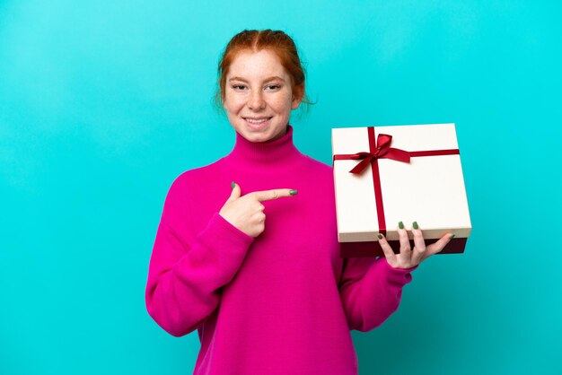 Jovem mulher avermelhada caucasiana segurando um presente isolado em fundo azul e apontando-o