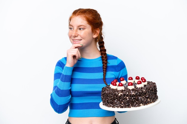 Jovem mulher avermelhada segurando bolo de aniversário isolado no fundo branco, olhando para o lado e sorrindo