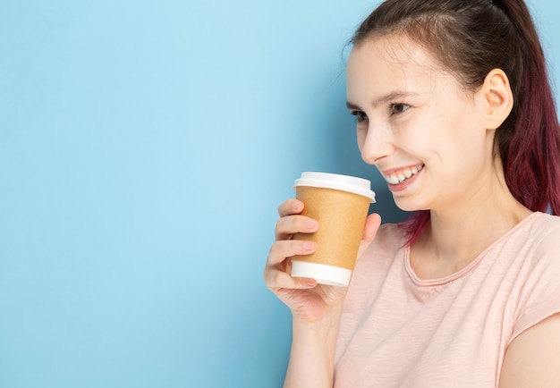 Jovem mulher bebe café de copo de papel e sorri sobre fundo azul