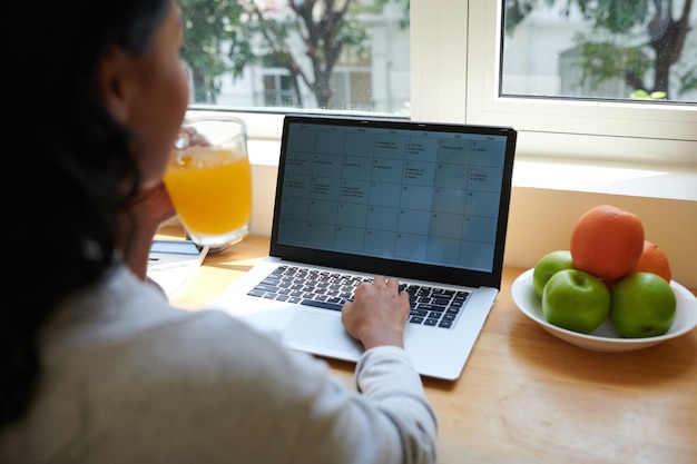 Jovem mulher bebendo suco e verificando calendário e agenda na tela do laptop