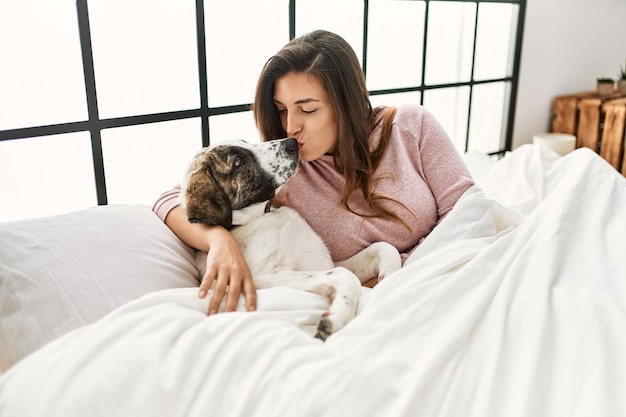 Jovem mulher beijando e abraçando cachorro sentado na cama no quarto