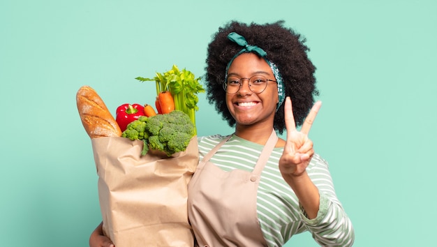 Jovem mulher bonita afro se sentindo feliz, relaxada e satisfeita, mostrando aprovação com um gesto de ok, sorrindo e segurando uma sacola de legumes