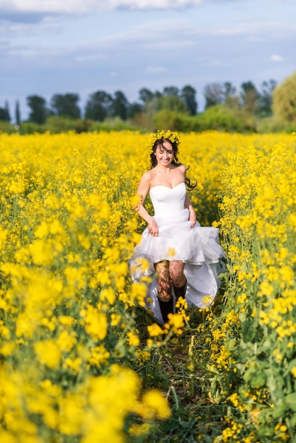 Jovem mulher bonita atravessa um campo com flores amarelas A noiva em um longo vestido branco e botas em um campo de colza