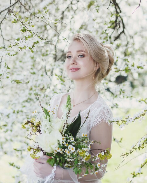 Jovem mulher bonita bonita posando em vestido de noiva de luxo contra flores em um dia ensolarado de verão. Retrato sensual da moda