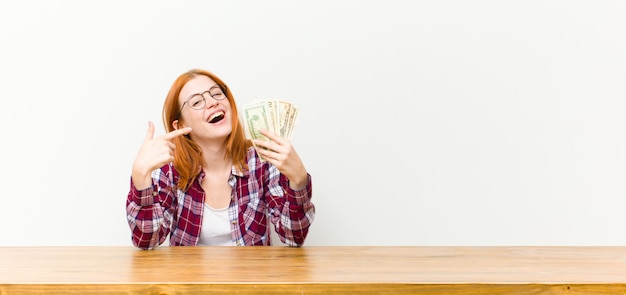 Jovem mulher bonita cabeça vermelha na frente de uma mesa de madeira com notas de dólar