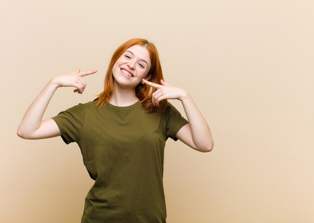 Jovem mulher bonita cabeça vermelha sorrindo confiante apontando para próprio sorriso largo, atitude positiva, relaxada, satisfeita