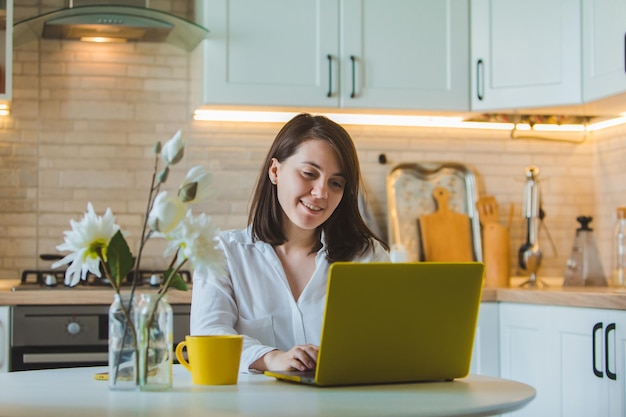 Jovem mulher bonita caucasiana falando ao telefone sentada em frente ao laptop na cozinha