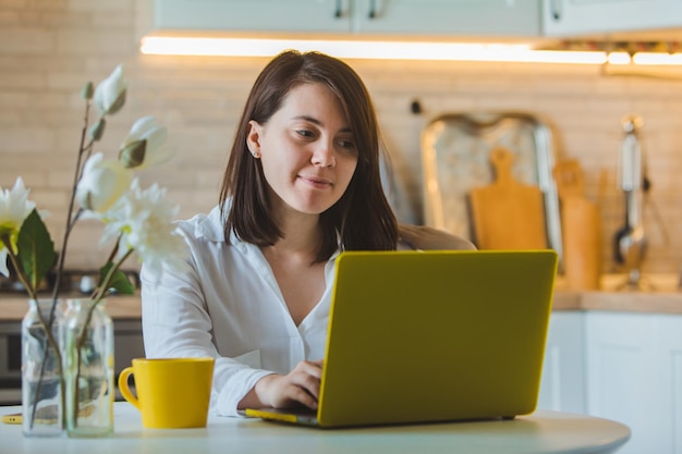 Jovem mulher bonita caucasiana falando ao telefone sentado na frente do laptop na cozinha. dona de casa de negócios. tomando café da caneca amarela