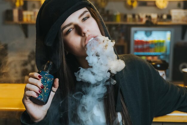 Foto jovem mulher bonita com boné fuma um cigarro eletrônico na loja de vape em close-up no estilo hip-hop