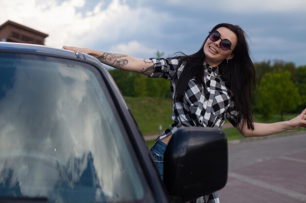 Jovem mulher bonita com cabelo comprido e camisa comprida na cabeça está de pé perto de um carro preto.