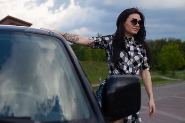 Jovem mulher bonita com cabelo comprido e cueca jeans na cabeça está de pé perto de um carro preto.