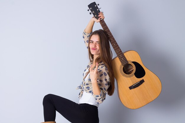 Jovem mulher bonita com cabelo comprido na blusa bege e calça preta segurando uma guitarra e mostrando ROCK em fundo cinza no estúdio