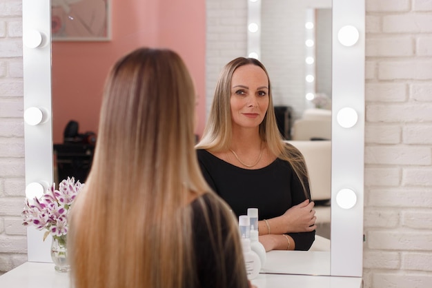 Jovem mulher bonita com cabelo loiro longa e saudável se olhando no espelho em um salão de cabeleireiro