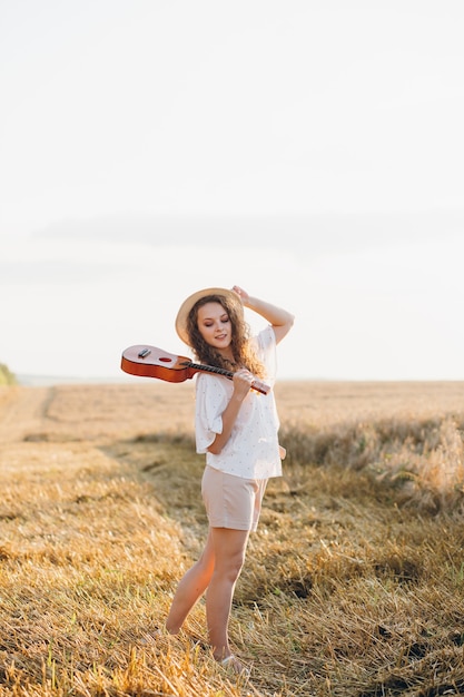 Jovem mulher bonita com cabelo longo encaracolado em shorts, um chapéu com uma guitarra posa em um campo de trigo no verão ao pôr do sol