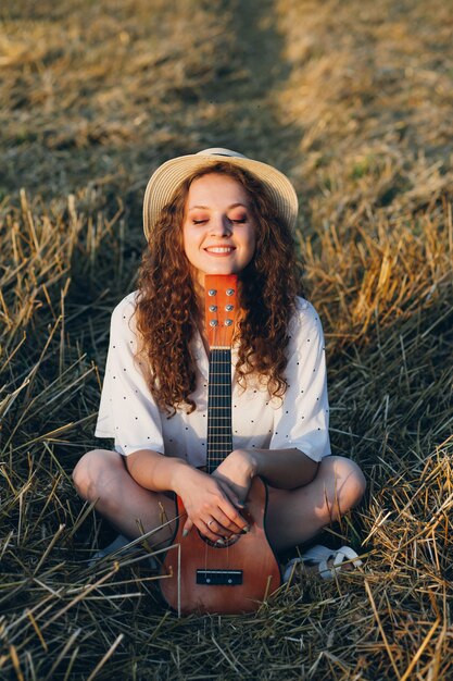 Jovem mulher bonita com cabelo longo encaracolado em shorts, um chapéu com uma guitarra posa em um campo de trigo no verão ao pôr do sol