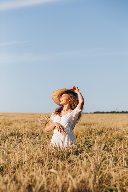 Jovem mulher bonita com cabelo longo encaracolado posa num campo de trigo no verão ao pôr do sol